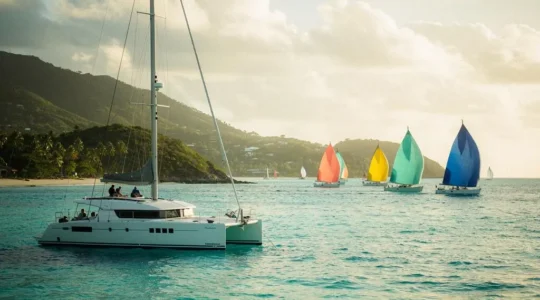 Un catamaran de spectateurs mouille dans une baie turquoise des Antilles pendant l'arrivée d'une flotte de voiliers de course au large